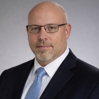 Black and white professional headshot of a bald man wearing glasses, dark blazer, and light blue tie against a neutral gray background